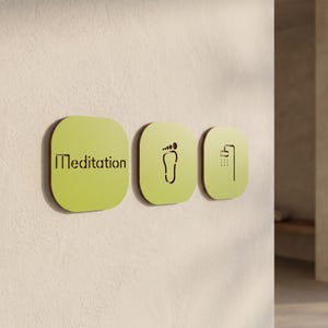 Può includere: Tre insegne quadrate verde chiaro con angoli arrotondati montate su una parete. La prima insegna presenta la parola Meditation in lettere nere ritagliate. La seconda mostra il contorno nero di un piede. La terza mostra l'icona di una doccia.