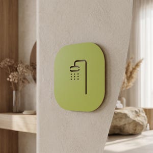 Puede incluir: Un letrero cuadrado de color verde lima con esquinas redondeadas que presenta un icono recortado de color marrón oscuro de un cabezal de ducha con gotas de agua. El letrero está montado en una pared texturizada de color blanco roto.