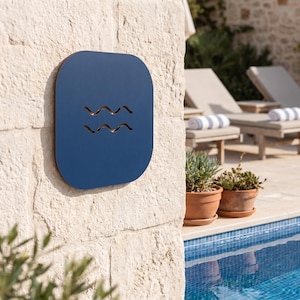 May include: A square, dark blue sign with rounded corners featuring two horizontal, wavy cut-out lines. The sign is mounted on a light-colored, textured stone wall next to a swimming pool with clear blue water and two potted plants nearby.