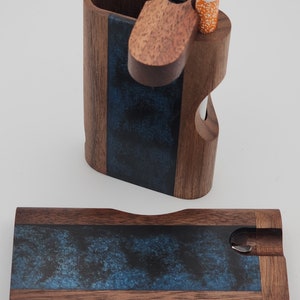 Peut inclure: Une boîte en bois avec un incrustation en résine bleue et un couvercle à charnière. Le couvercle a un petit trou pour une cigarette et une fente pour un briquet. La boîte est fermée dans la deuxième image.