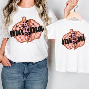 May include: Two white t-shirts with a pumpkin design. The larger shirt says "mama" and the smaller shirt says "mini". Both pumpkins are orange with pink and black checkered patterns and a lightning bolt with a leopard print.