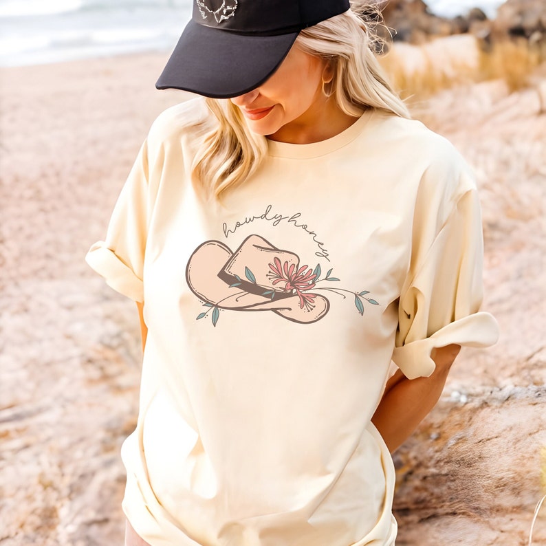 K&ouml;nnte beinhalten: Ein hellgelbes T-Shirt mit einem Aufdruck eines Cowboyhuts mit einer Blume und dem Text "howdy honey".