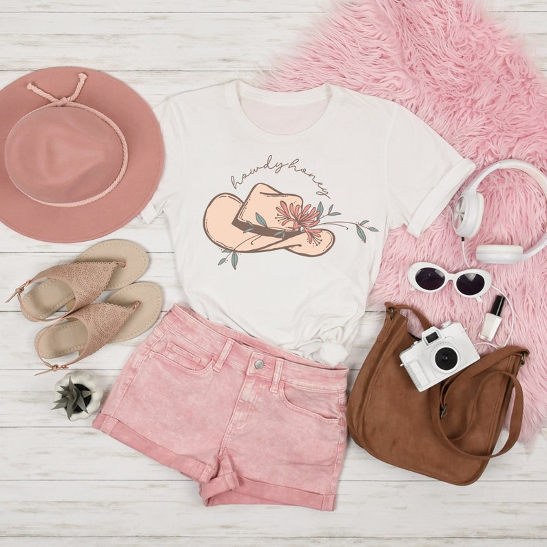 K&ouml;nnte beinhalten: Ein wei&szlig;es T-Shirt mit einem rosafarbenen Cowboyhut und einem floralen Design. Der Text "howdy honey" ist in einer Schreibschrift &uuml;ber dem Hut geschrieben. Das Shirt liegt auf einer wei&szlig;en Holzoberfl&auml;che mit rosafarbenen Jeans-Shorts, einer braunen Ledertasche, einem rosafarbenen Hut, Sandalen, Sonnenbrillen und Kopfh&ouml;rern.