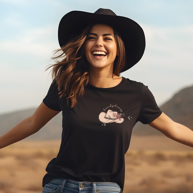 K&ouml;nnte beinhalten: Ein schwarzes T-Shirt mit einem rosafarbenen Cowboyhut und einem Blumendesign. Der Text "howdy fancy" ist auf dem Shirt geschrieben.