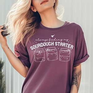 May include: A purple t-shirt with the text "always feeding my sourdough starter" and a graphic of three jars with lids and ribbons.