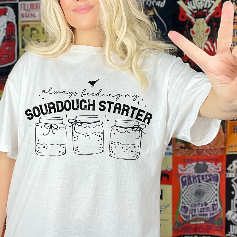 May include: White t-shirt with black text that reads "always feeding my sourdough starter" and a graphic of three jars with lids. Each jar has a small bird on top and a swirl design inside.