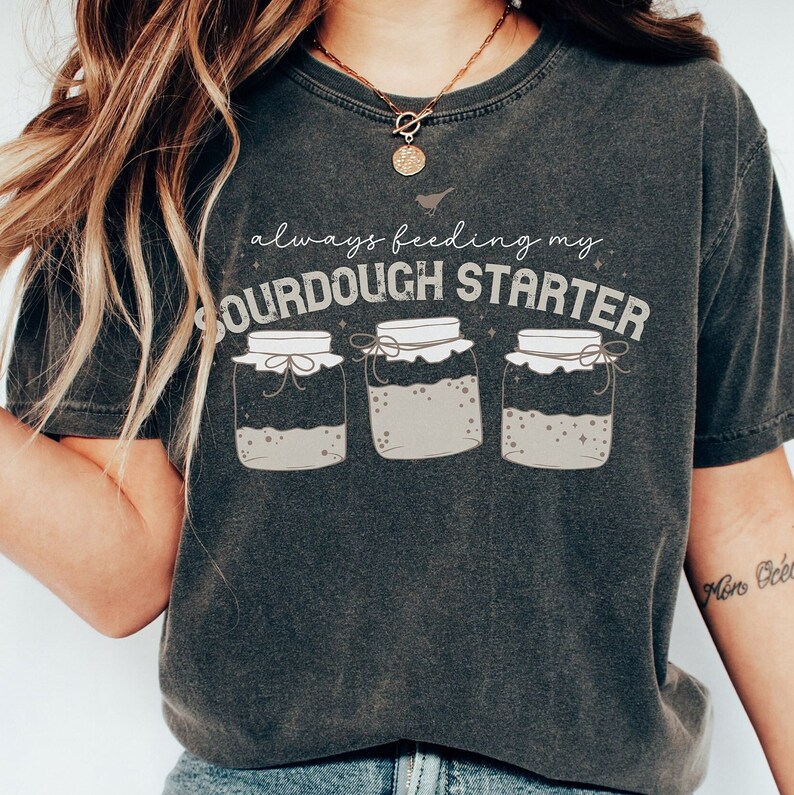 May include: A dark gray t-shirt with the text "always feeding my sourdough starter" and three illustrations of jars with sourdough starter inside.