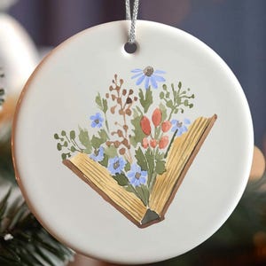 May include: A round, white ceramic Christmas ornament with a watercolor illustration of an open book filled with colorful flowers. The ornament hangs from a silver string and is displayed on a Christmas tree.