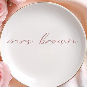 May include: A white ceramic plate with a gold rim, inscribed with "mrs. brown" in a delicate script. The plate is surrounded by pink roses, green leaves, and red heart-shaped candies, all arranged on a pink fabric background.