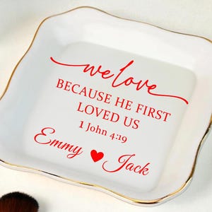 May include: A white ceramic trinket dish with a gold rim. Red script reads "we love BECAUSE HE FIRST LOVED US 1 John 4:19 Emmy ❤️ Jack". The dish has a unique, irregular shape and is surrounded by makeup brushes and a decorative ornament.