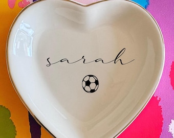 May include: A heart-shaped, white ceramic trinket dish with a gold rim. The dish features the name "sarah" in cursive and a small soccer ball graphic. The background is a colorful abstract design.