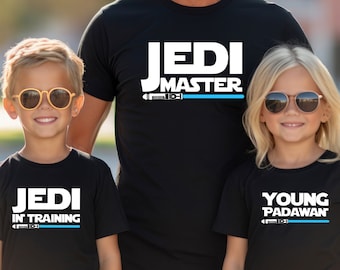 Puede incluir: Tres personas con camisetas negras con texto blanco y un gráfico de sable de luz azul. Las camisetas dicen "Jedi Master", "Jedi in Training" y "Young Padawan".