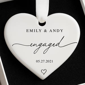 K&ouml;nnte beinhalten: Ein wei&szlig;er herzf&ouml;rmiger Schmuckanh&auml;nger mit dem Text "EMILY & ANDY engaged 05.27.2021" und einem Herzsymbol. Der Anh&auml;nger h&auml;ngt an einem wei&szlig;en Band und wird in einer wei&szlig;en Schachtel mit schwarzem Innenraum pr&auml;sentiert.