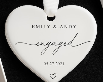 May include: A white heart-shaped ornament with the text "EMILY & ANDY engaged 05.27.2021" and a heart symbol. The ornament hangs from a white ribbon and is presented in a white box with a black interior.