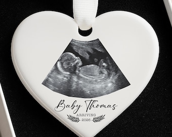 May include: A white heart-shaped ornament with a ribbon, featuring a sonogram image. The ornament is inscribed with "Baby Thomas ARRIVING 2026" in elegant script, presented in a gift box with a black interior.