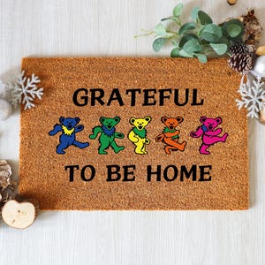 May include: A rectangular doormat made of natural coir fiber. The mat features the words "GRATEFUL TO BE HOME" in black, with colorful dancing bear graphics in blue, green, yellow, orange, and pink.