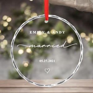 K&ouml;nnte beinhalten: Ein klarer, facettierter Glasornament mit den Worten "EMILY & ANDY" und "married" in wei&szlig;er Schrift. Das Datum "05.27.2021" und ein Herz sind ebenfalls abgebildet. Ein rotes Band ist zum Aufh&auml;ngen befestigt.