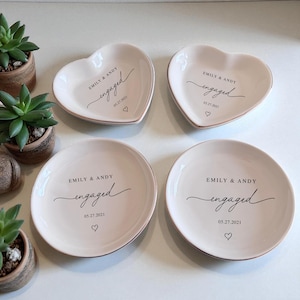 May include: Four off-white ring dishes, two heart-shaped and two round. Each dish has the names "EMILY & ANDY", the word "engaged", the date "05.27.2021", and a heart symbol in black script. The rims have a subtle gold trim.