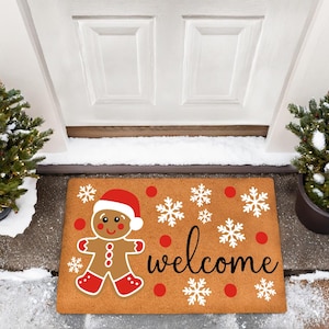 May include: A rectangular doormat featuring a gingerbread man in a Santa hat and the word "welcome" in black script. The mat has red polka dots and white snowflakes on a brown background. Two small evergreen trees flank the door.