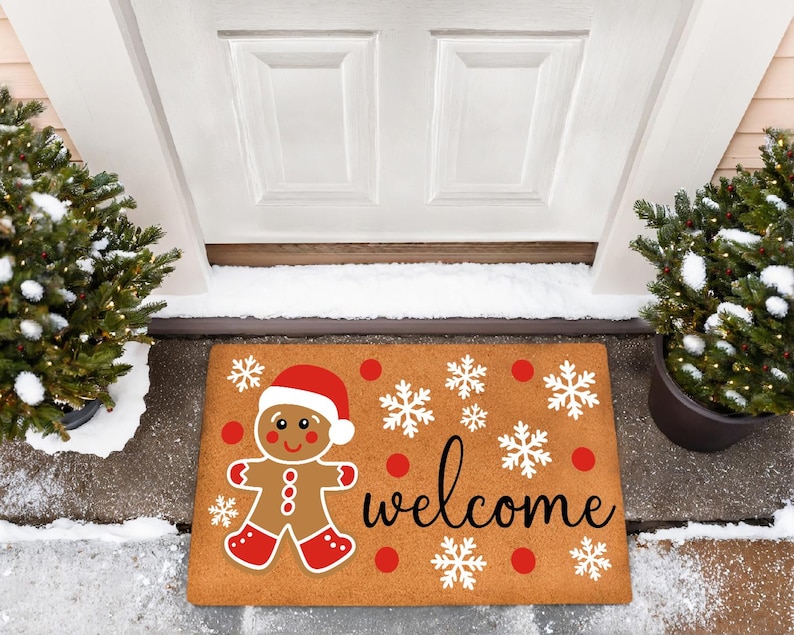 May include: A rectangular doormat featuring a gingerbread man in a Santa hat and the word "welcome" in black script. The mat has red polka dots and white snowflakes on a brown background. Two small evergreen trees flank the door.