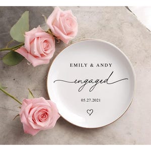 May include: A white ceramic ring dish with a gold rim, inscribed with "EMILY & ANDY engaged 05.27.2021" in black script, and a heart. The dish is set amongst three pink roses with green foliage.
