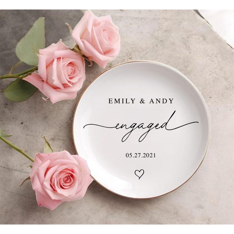 May include: A white ceramic ring dish with a gold rim, inscribed with "EMILY & ANDY engaged 05.27.2021" in black script, and a heart. The dish is set amongst three pink roses with green foliage.