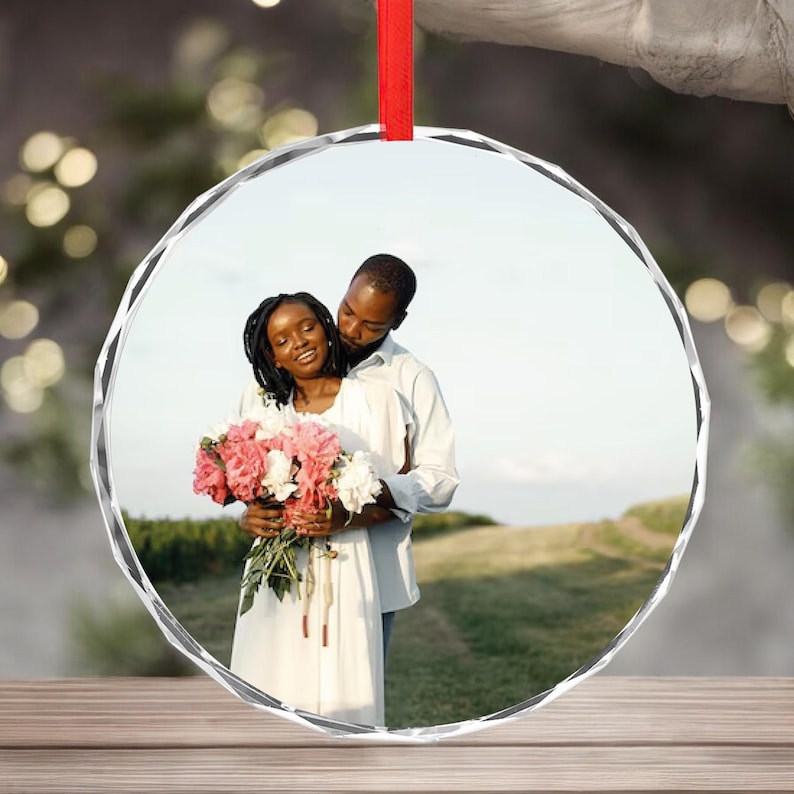 May include: A clear, round glass ornament with a red ribbon. The ornament features a photograph of a couple embracing, the woman holding a bouquet of pink and white flowers. The background shows a field and sky.