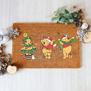 May include: A rectangular brown doormat featuring three Winnie the Pooh characters in Christmas attire. One is dressed as a Christmas tree, another is wrapped in lights, and the third holds a green scarf that says "Oh Joy".