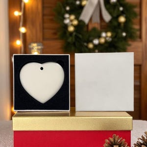 K&ouml;nnte beinhalten: Ein herzf&ouml;rmiger, cremefarbener Ornament mit einem kleinen Loch zum Aufh&auml;ngen, pr&auml;sentiert in einer schwarz gef&uuml;tterten Box. Die Box steht auf einer rot-goldenen Geschenkbox, mit Tannenzapfen im Vordergrund. Ein Weihnachtskranz ist im Hintergrund.