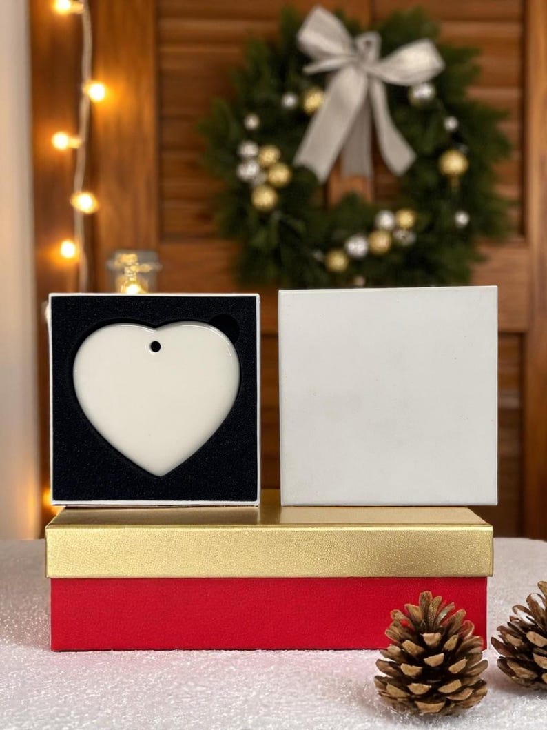 K&ouml;nnte beinhalten: Ein herzf&ouml;rmiger, cremefarbener Ornament mit einem kleinen Loch zum Aufh&auml;ngen, pr&auml;sentiert in einer schwarz gef&uuml;tterten Box. Die Box steht auf einer rot-goldenen Geschenkbox, mit Tannenzapfen im Vordergrund. Ein Weihnachtskranz ist im Hintergrund.