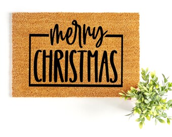 May include: A rectangular doormat made of natural coir fiber. The mat features the words "merry CHRISTMAS" in a black, bold font within a black rectangular border. A small green plant is in the lower right corner, set against a white background.