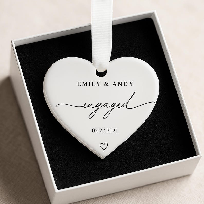 K&ouml;nnte beinhalten: Ein wei&szlig;er herzf&ouml;rmiger Schmuckanh&auml;nger mit dem Text "EMILY & ANDY engaged 05.27.2021" und einem Herzsymbol. Der Anh&auml;nger h&auml;ngt an einem wei&szlig;en Band und wird in einer wei&szlig;en Schachtel mit schwarzem Innenraum pr&auml;sentiert.