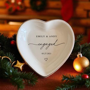 May include: A heart-shaped, white ceramic dish with a gold rim. The dish is inscribed with "EMILY & ANDY" above the word "engaged" and the date "05.27.2021". A small heart symbol is below the date. Festive holiday decorations surround the dish.
