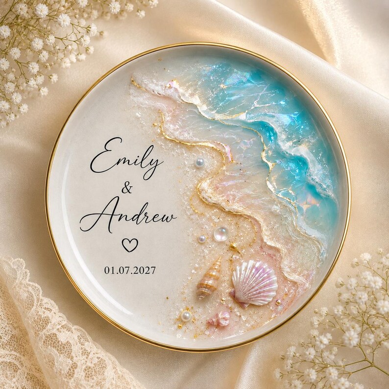 May include: A round ceramic ring dish with a gold rim featuring a beach-themed resin design with blue and white waves, sand, small seashells, and pearls. The text reads Emily & Andrew with a heart symbol and the date 01.07.2027 in black script.
