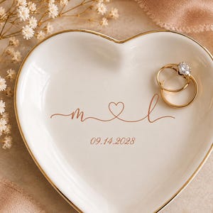 May include: A white heart-shaped ceramic ring dish with a gold-painted rim. The center features copper-colored cursive letters M and L connected by a small heart, with the date 09.14.2028 printed below. Two gold-toned metal rings rest on the dish.