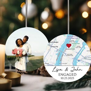 May include: Round ceramic ornament with two sides. One side features a couple holding flowers, the other a map of Manhattan with a heart marking a location. Text reads "Lisa & John ENGAGED 10.25.2025".