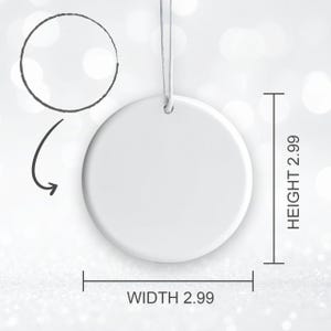 May include: A white, round ornament with a string for hanging. The ornament is 2.99 inches in width and 2.99 inches in height. A black arrow and circle are drawn above the ornament. The background is a soft, blurred white.