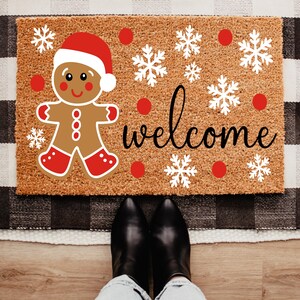 May include: A rectangular doormat featuring a gingerbread man in a Santa hat, snowflakes, and the word "welcome" in black script. The mat is brown with a textured surface, and the design includes red and white accents.