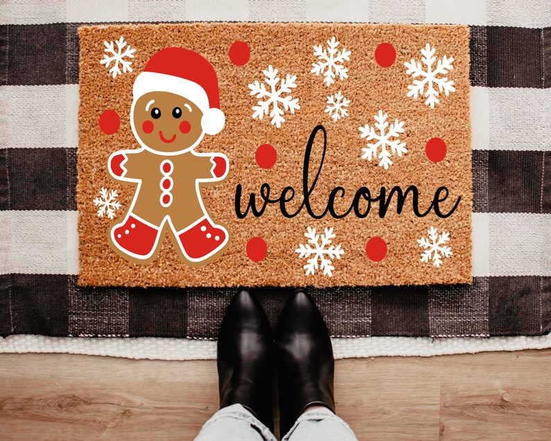 May include: A rectangular doormat featuring a gingerbread man in a Santa hat, snowflakes, and the word "welcome" in black script. The mat is brown with a textured surface, and the design includes red and white accents.