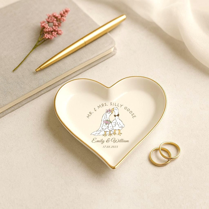 Puede incluir: Un joyero de cer&aacute;mica en forma de coraz&oacute;n, de color crema con borde dorado. El plato presenta una ilustraci&oacute;n caprichosa de dos gansos con atuendo de boda, con el texto "Mr. & Mrs. Silly Goose" y una fecha personalizada. Dos anillos de oro est&aacute;n junto al plato.