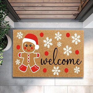 May include: A rectangular doormat featuring a gingerbread man in a Santa hat, snowflakes, and the word "welcome". The mat is brown with red and white details, ideal for the festive period.