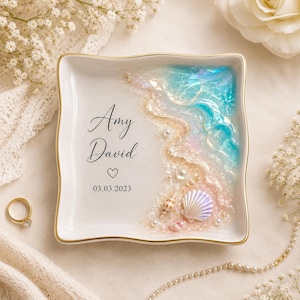May include: A square white ceramic ring dish with gold trim featuring a resin ocean wave design on the right side. The dish is personalized with the names Amy and David, a heart symbol, and the date 03.03.2023 in black script and sans-serif text.