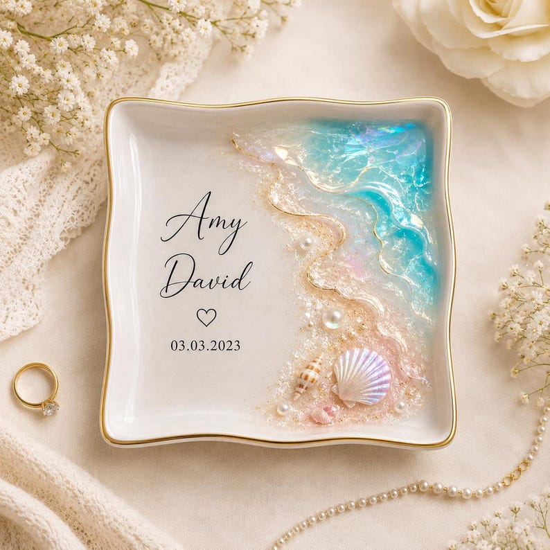 May include: A square white ceramic ring dish with gold trim featuring a resin ocean wave design on the right side. The dish is personalized with the names Amy and David, a heart symbol, and the date 03.03.2023 in black script and sans-serif text.