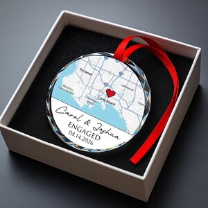 May include: A clear, faceted glass ornament with a map of Long Beach, CA, and a red heart marking a location. The ornament has a red ribbon and text that reads "Carol & Joshua ENGAGED 08.14.2026". It is presented in a white box with a black interior.