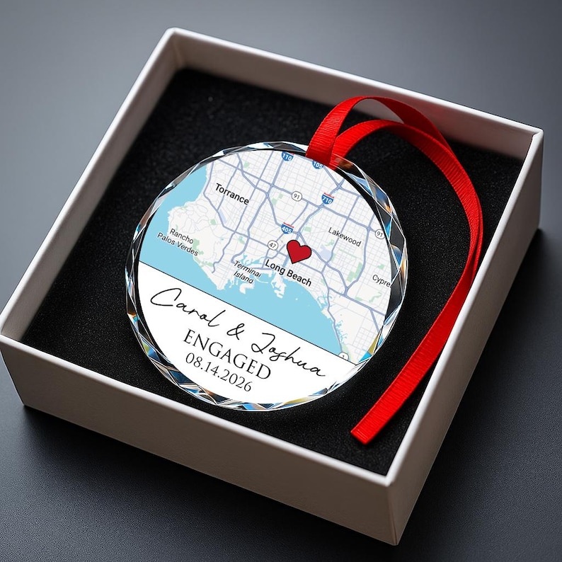 May include: A clear, faceted glass ornament with a map of Long Beach, CA, and a red heart marking a location. The ornament has a red ribbon and text that reads "Carol & Joshua ENGAGED 08.14.2026". It is presented in a white box with a black interior.