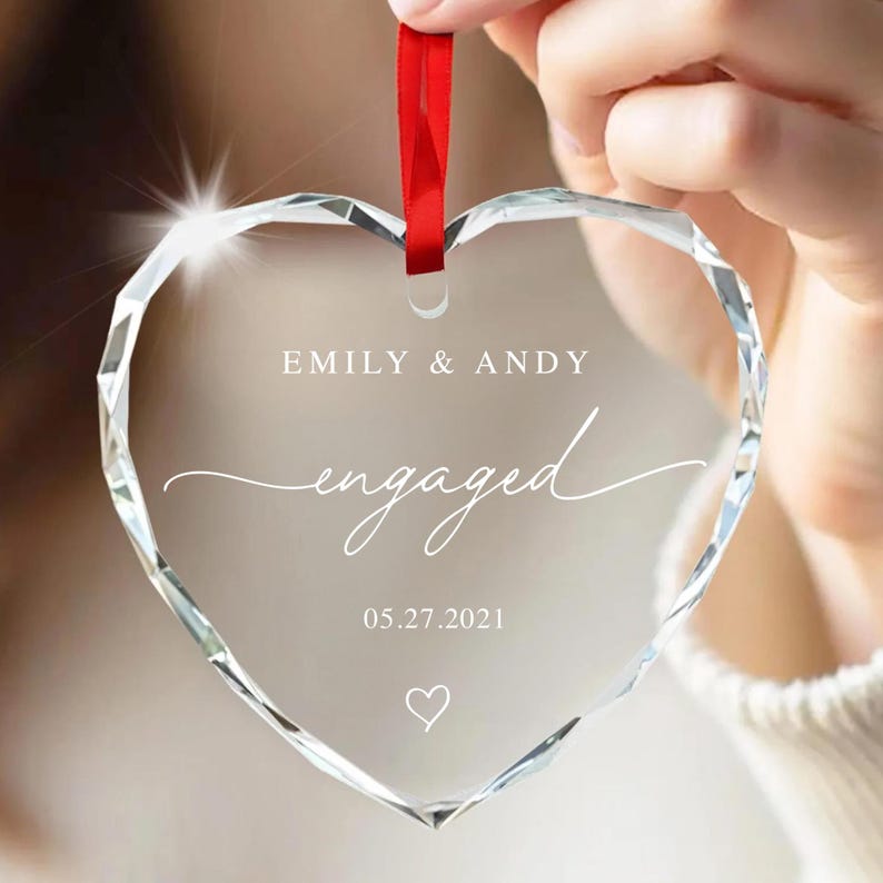 K&ouml;nnte beinhalten: Ein klarer, herzf&ouml;rmiger Ornament mit facettiertem Design. Das Ornament tr&auml;gt die Worte "Emily & Andy" und "engaged" in wei&szlig;er Schrift, zusammen mit dem Datum 05.27.2021 und einem Herzsymbol. Ein rotes Band ist zum Aufh&auml;ngen befestigt.