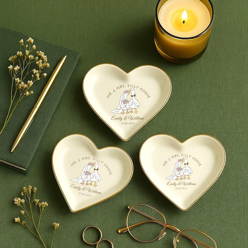 Puede incluir: Tres platos para anillos en forma de coraz&oacute;n con bordes dorados. Cada plato presenta una caricatura de dos gansos con atuendo de boda, el texto "MR. & MRS. SILLY GOOSE" y los nombres "Emily & William" con una fecha. Tambi&eacute;n se ven una vela encendida y un bol&iacute;grafo.