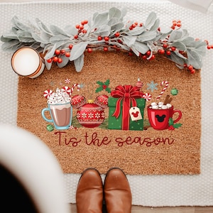 May include: A festive doormat with a Christmas theme. It displays a hot chocolate mug, baubles, a wrapped gift, and a red mug with marshmallows. The text "Tis the season" is written across the bottom. A candle and decorative garland are also present.