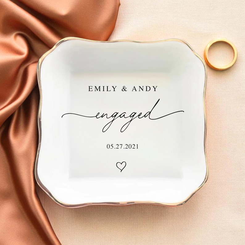 May include: A white ceramic ring dish with a gold rim and scalloped edges. The dish is inscribed with "EMILY & ANDY engaged 05.27.2021" and a heart symbol. A gold ring rests on the side.