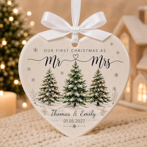 May include: A heart-shaped ceramic ornament featuring a white ribbon bow at the top. The surface displays three illustrated pine trees, snowflakes, and the text "OUR FIRST CHRISTMAS AS Mr & Mrs", "Thomas & Emily", and the date "01.05.2027".